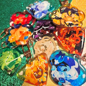May include: A selection of vibrant acrylic keyrings showcasing diverse character designs. The keyrings are in various shapes, including hearts and flower-like forms, with gold-coloured metal clasps. The designs feature shades of red, orange, blue, and green.