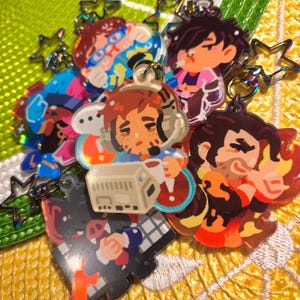 Könnte beinhalten: Eine Sammlung farbenfroher Acryl-Schlüsselanhänger mit Cartoon-Illustrationen verschiedener Charaktere. Die Schlüsselanhänger haben eine glänzende Oberfläche und sind an Metall-Schlüsselringen befestigt. Die Designs umfassen Figuren mit Brillen, Kopfhörern und anderem Zubehör.