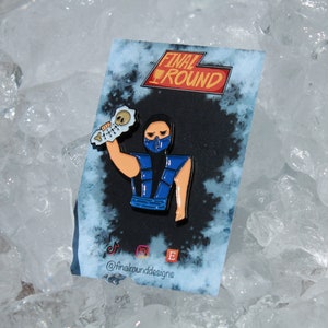 以下が含まれることがあります： ビデオゲーム「Mortal Kombat」のキャラクターをフィーチャーした青と黒のエナメルピン。キャラクターは骨を持っており、青いマスクをしています。ピンには上部に「Final Round」、下部に「@finalrounddesigns」と書いてあります。