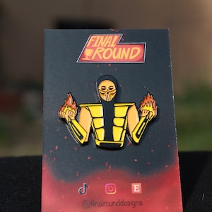 Könnte beinhalten: Eine gelbe und schwarze Emaille-Anstecknadel mit einer Figur aus dem Videospiel Mortal Kombat. Die Figur hält in jeder Hand zwei brennende Gegenstände. Die Anstecknadel befindet sich auf einem schwarzen Hintergrund mit dem Text "FINAL ROUND" in Rot und Weiß. Der Text "@finalrounddesigns" ist ebenfalls auf der Karte zu sehen.
