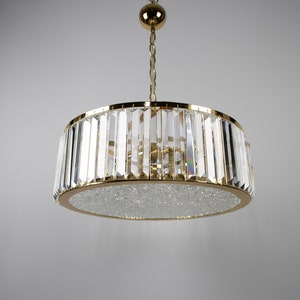 Pu&ograve; includere: Un lampadario dorato con un paralume rotondo in cristallo. Il paralume &egrave; fatto di molti piccoli cristalli trasparenti disposti in un motivo geometrico.