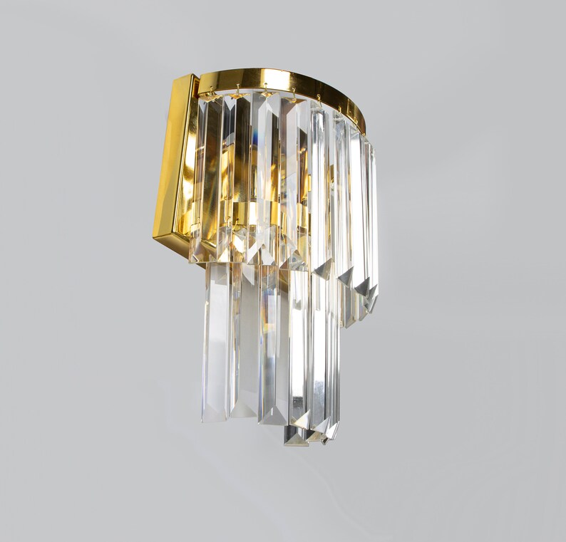 May include: A gold wall sconce with cascading crystal prisms. The sconce has a curved design and is mounted on a wall.
