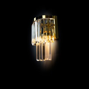 May include: A gold wall sconce with clear crystal prisms hanging down. The light is on and illuminating the crystals.