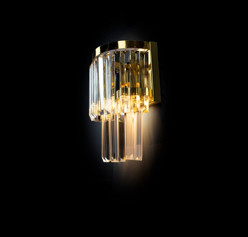 May include: A gold wall sconce with clear crystal prisms hanging down. The light is on and illuminating the crystals.