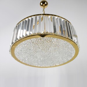 Pu&ograve; includere: Un lampadario dorato con una base rotonda ricoperta di cristalli e un anello di prismi di cristallo trasparente attorno alla parte superiore.