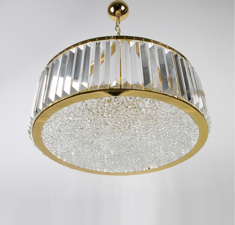 Pu&ograve; includere: Un lampadario dorato con una base rotonda ricoperta di cristalli e un anello di prismi di cristallo trasparente attorno alla parte superiore.