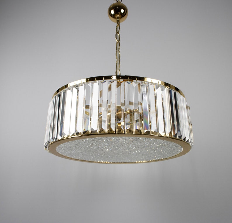 Pu&ograve; includere: Un lampadario dorato con un paralume rotondo in cristallo. Il paralume &egrave; fatto di molti piccoli cristalli trasparenti disposti in un motivo geometrico.