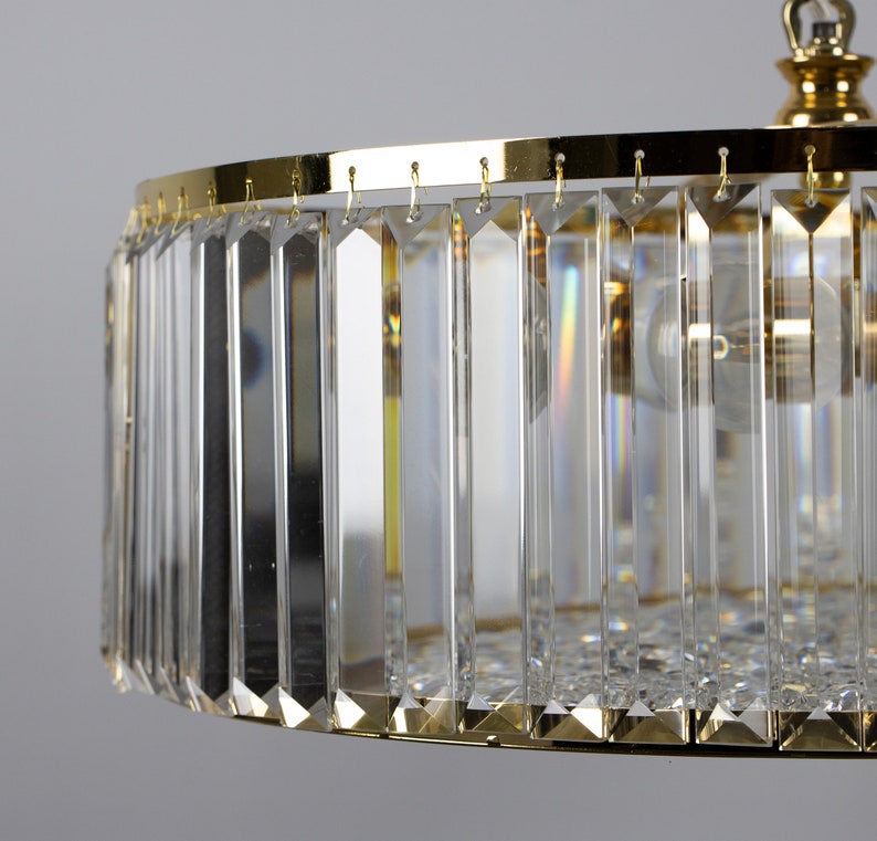 Pu&ograve; includere: Un lampadario dorato con un paralume rotondo in cristallo. Il paralume &egrave; composto da molti prismi di cristallo verticali.