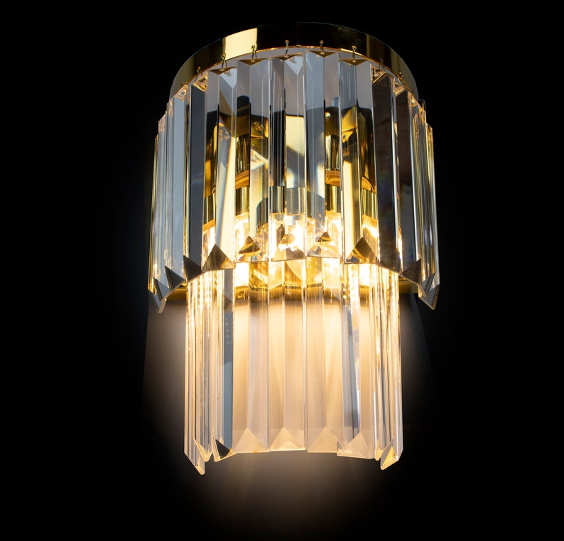 May include: A gold-toned wall sconce with a crystal design. The sconce has two tiers of crystal prisms that hang down from a circular base.
