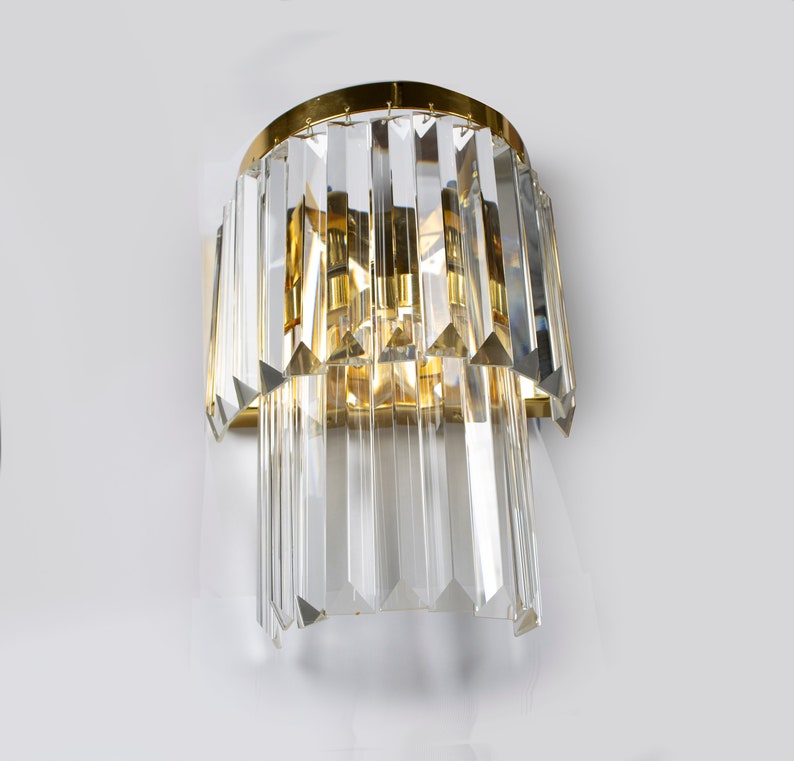 May include: A gold-toned wall sconce with a cascading design of clear crystal prisms. The sconce has a cylindrical base and a modern, minimalist style.