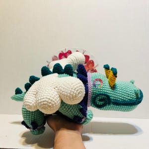 May include: Crocheted dinosaur toy in shades of teal, white, and purple. The handmade plushie features a detailed design with spikes, a rainbow emblem, and yellow accents. A unique, handcrafted item.