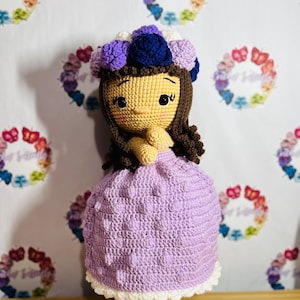May include: A handmade crochet doll with a light brown face, dark brown hair, and a purple dress. The doll wears a flower crown with purple, blue, and white flowers. The doll is displayed on a glass base.