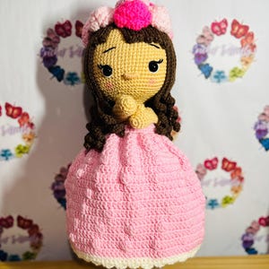 May include: A handmade crochet doll with a light brown face, dark brown hair, and a pink dress. The doll has a pink and light pink floral crown. The doll is standing on a clear glass base.
