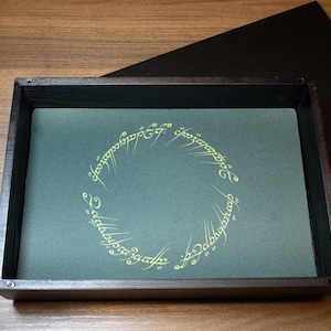 May include: A wooden box with a teal interior featuring gold lettering in a circular pattern. A black lid with gold text and a cartoon image is resting on the side. The box is on a wooden surface.