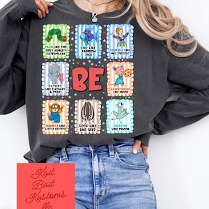 Peut inclure: Sweat-shirt gris foncé avec des carrés graphiques colorés représentant des personnages et des phrases comme "Calme comme la très grosse chenille" et "Soyez". Le t-shirt a un col rond et des manches longues. Le nom de la marque "Kait Bait Kustoms llc" est imprimé sur un carré rouge.