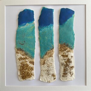 May include: Abstract artwork in a white frame, comprising three vertical rectangular forms. Each form displays a colour gradient: dark blue at the top, fading to turquoise, then beige, and finally white at the base. The piece suggests a coastal theme, reminiscent of sea and sand.