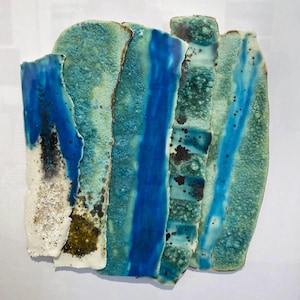 May include: Abstract ceramic artwork with a layered design. The piece features vertical bands of varying widths and textures in shades of blue, turquoise, and white, with dark speckles. The artwork has a rough, organic appearance.