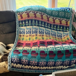 May include: A colorful crocheted throw blanket with a winter theme. The blanket features a repeating pattern of snowflakes, mittens, and reindeer. The text "Snow Snow Snow Day Day Day Day Day Day Day Day" is woven into the blanket.