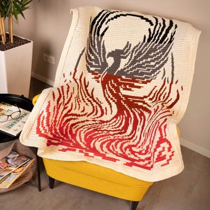 Peut inclure: Une couverture en crochet représentant un phénix s'élevant des flammes. Le phénix est représenté dans des tons de gris, de noir et de rouge, tandis que les flammes sont un dégradé de rouge et d'orange. La couverture est réalisée avec un fil blanc et présente un motif texturé en crochet.