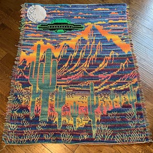 Peut inclure: Une tapisserie en crochet color&eacute;e repr&eacute;sentant un paysage d&eacute;sertique avec de grands cactus et une soucoupe volante dans le ciel. La tapisserie a une bordure frang&eacute;e.