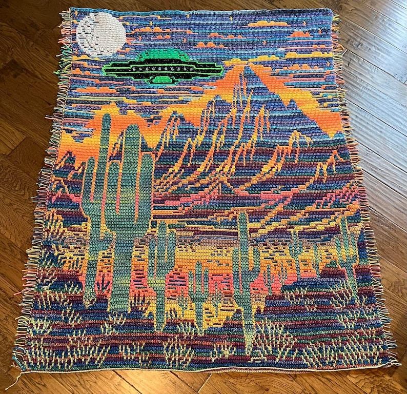 Peut inclure: Une tapisserie en crochet color&eacute;e repr&eacute;sentant un paysage d&eacute;sertique avec de grands cactus et une soucoupe volante dans le ciel. La tapisserie a une bordure frang&eacute;e.
