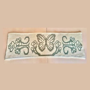 Puede incluir: Camino de mesa rectangular de ganchillo blanco roto con un diseño central de mariposa en tonos grises y verdes. Motivos florales y de volutas flanquean la mariposa. El camino de mesa es probablemente hecho a mano y añade un toque natural.