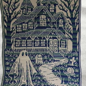Op de afbeelding: Een gehaakte deken met een spookhuis sc&egrave;ne in groen, blauw en beige tinten. Het ontwerp omvat een spookhuis, een geest, een kerkhof en bomen, waardoor een Halloween- of gothic thema ontstaat.