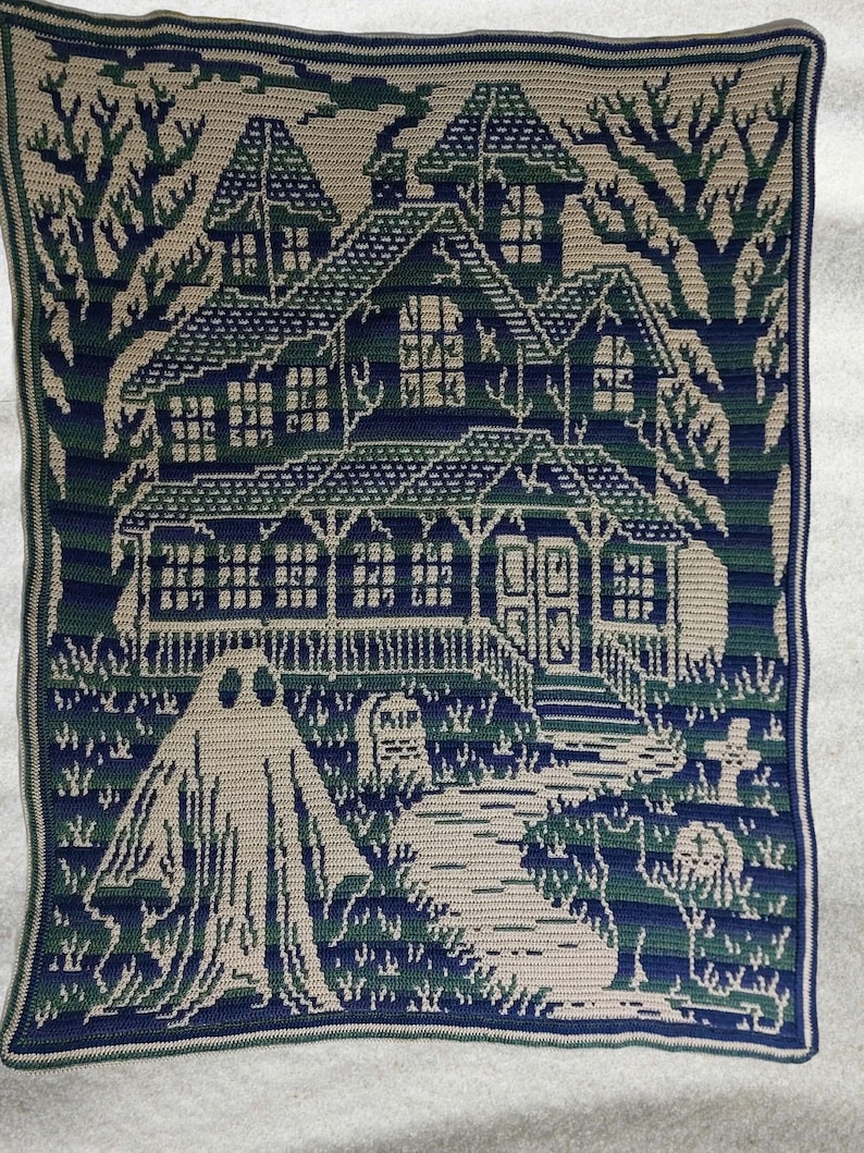Op de afbeelding: Een gehaakte deken met een spookhuis sc&egrave;ne in groen, blauw en beige tinten. Het ontwerp omvat een spookhuis, een geest, een kerkhof en bomen, waardoor een Halloween- of gothic thema ontstaat.