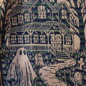 Op de afbeelding: Een gehaakte deken met een spookachtig spookhuis. Het ontwerp omvat een huis met meerdere ramen, een geest en een begraafplaats, allemaal in tinten blauw, groen en cr&egrave;me. De deken is waarschijnlijk handgemaakt en geschikt voor Halloween.
