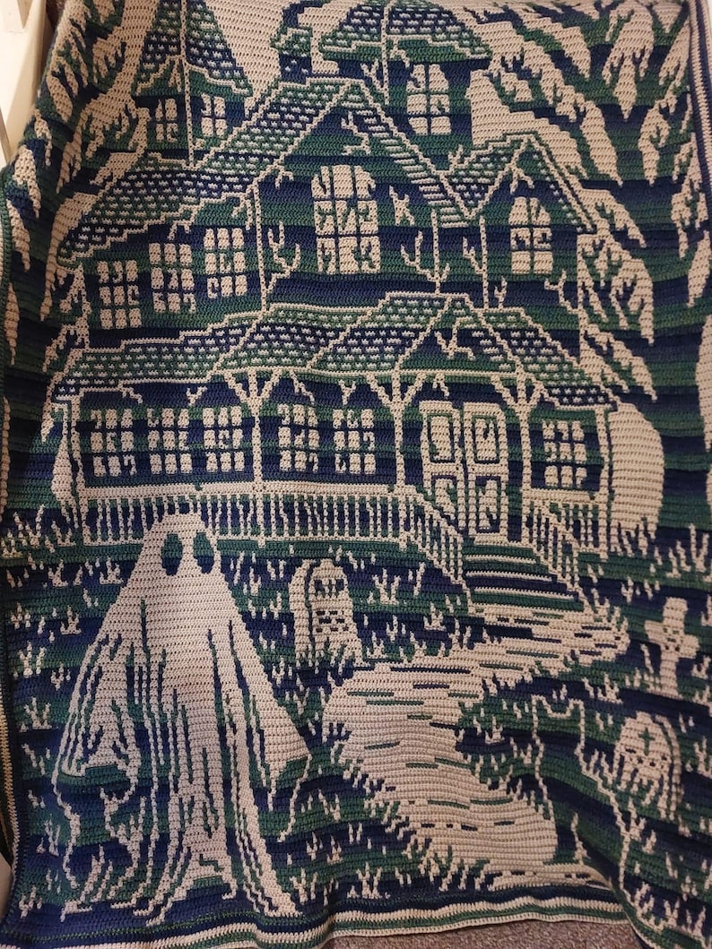 Op de afbeelding: Een gehaakte deken met een spookachtig spookhuis. Het ontwerp omvat een huis met meerdere ramen, een geest en een begraafplaats, allemaal in tinten blauw, groen en cr&egrave;me. De deken is waarschijnlijk handgemaakt en geschikt voor Halloween.