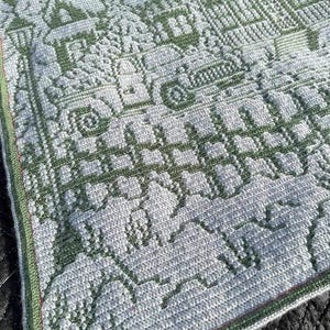 May include: A green and white crocheted blanket with a winter scene design. The blanket features houses, trees, and snowflakes.