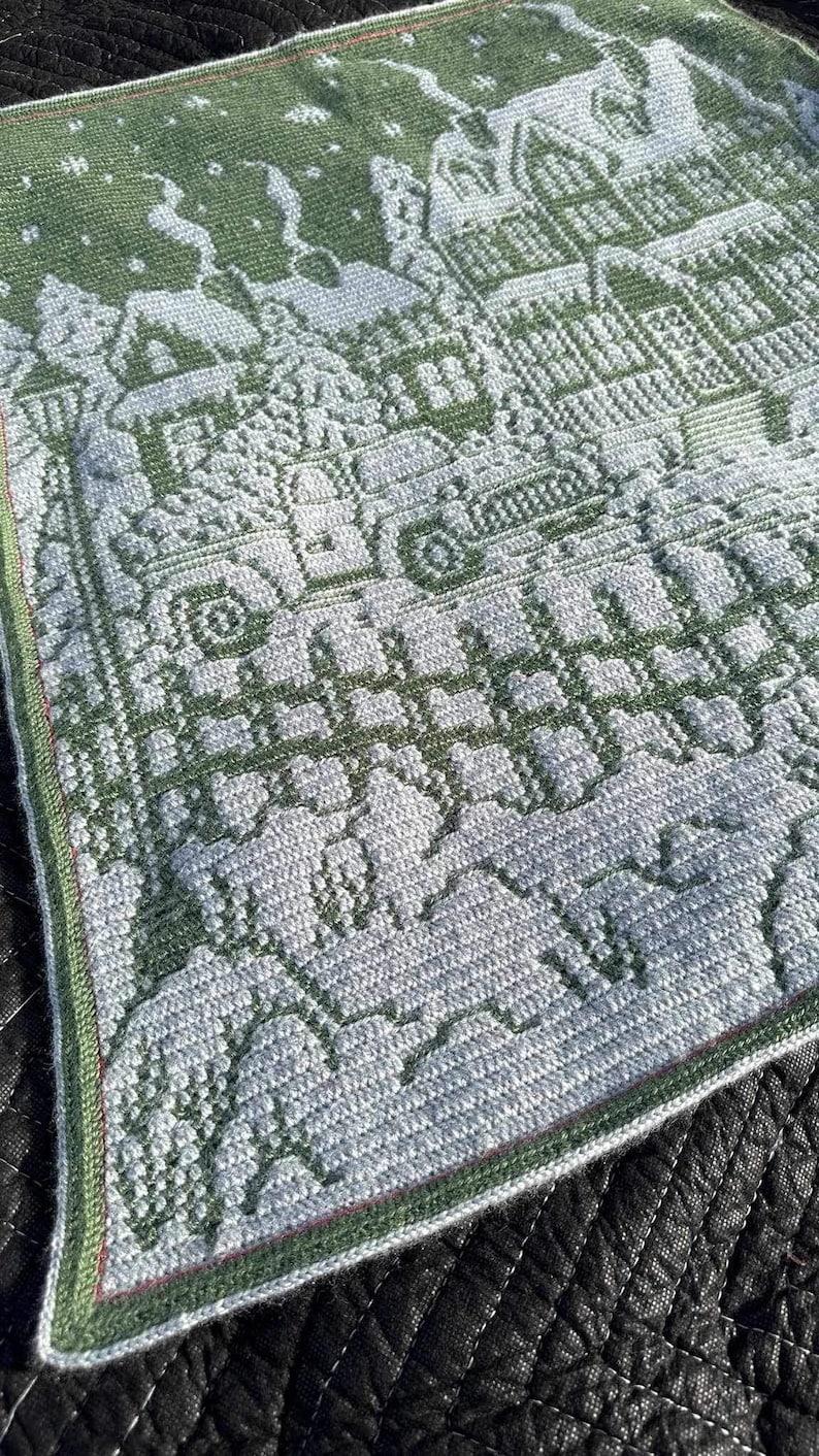 May include: A green and white crocheted blanket with a winter scene design. The blanket features houses, trees, and snowflakes.