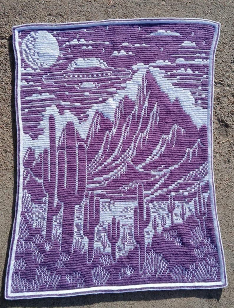Peut inclure: Une couverture en crochet de couleur violette et blanche avec un motif de paysage d&eacute;sertique. Le motif repr&eacute;sente des montagnes, une lune et des cactus.