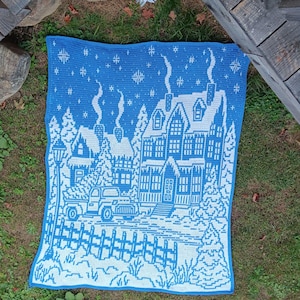 May include: A blue and white crocheted blanket with a winter scene of a house, trees, a car, and a fence. The house has a snowy roof and icicles hanging from the eaves. The blanket is made with a simple stitch pattern and has a border of blue yarn.