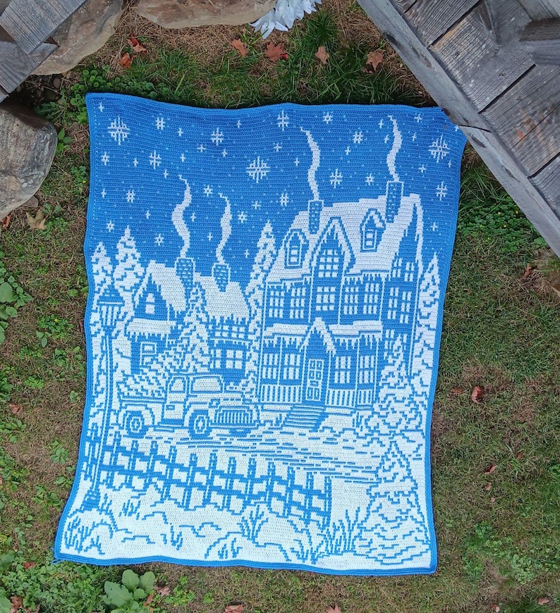 May include: A blue and white crocheted blanket with a winter scene of a house, trees, a car, and a fence. The house has a snowy roof and icicles hanging from the eaves. The blanket is made with a simple stitch pattern and has a border of blue yarn.