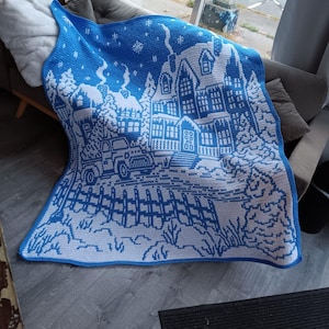 May include: A white and blue crocheted throw blanket with a winter scene featuring a house, trees, and a fence. The blanket is made with a chunky yarn and has a textured surface.