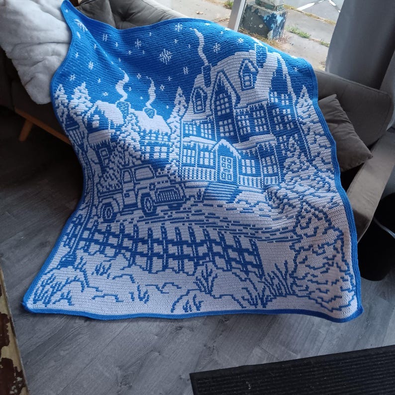May include: A white and blue crocheted throw blanket with a winter scene featuring a house, trees, and a fence. The blanket is made with a chunky yarn and has a textured surface.