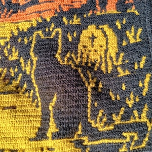 Op de afbeelding: Een close-up van een gehaakte textiel met een katten silhouet in geel tegen een donkerblauwe achtergrond. Het ontwerp bevat oranje en gele accenten, met een getextureerd uiterlijk. Het algemene thema is handgemaakt.