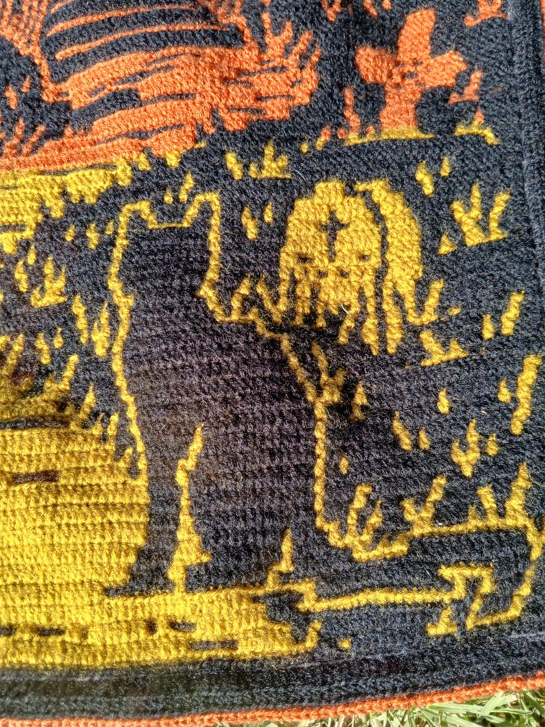 Op de afbeelding: Een close-up van een gehaakte textiel met een katten silhouet in geel tegen een donkerblauwe achtergrond. Het ontwerp bevat oranje en gele accenten, met een getextureerd uiterlijk. Het algemene thema is handgemaakt.