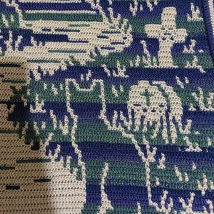 Op de afbeelding: Gehaakte deken met een landschapsontwerp met bomen, een kruis en een hond. De deken gebruikt tinten blauw, groen en beige, waardoor een getextureerd, handgemaakt uiterlijk ontstaat. Het ontwerp is ingewikkeld en gedetailleerd.