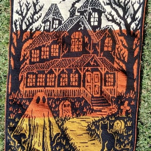 Op de afbeelding: Een Halloween-thema wandtapijt met een spookhuis met oranje en zwarte details. Het ontwerp omvat een geest, een zwarte kat en een pad dat naar het huis leidt. Het wandtapijt is geplaatst tegen een achtergrond van bomen en een bewolkte lucht.