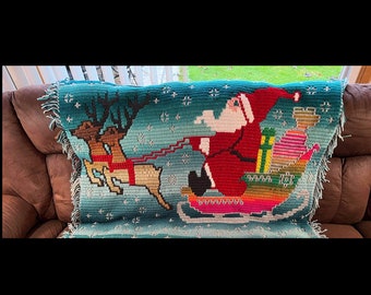 Pode incluir: Uma manta de crochê com uma representação colorida do Papai Noel em um trenó puxado por renas. O fundo é uma cena de neve azul e branca.