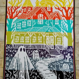 Op de afbeelding: Een kleurrijke gehaakte deken met een spookhuis-sc&egrave;ne. Het ontwerp omvat een geest, grafstenen en een pad dat naar een huis leidt. De deken gebruikt een verscheidenheid aan kleuren, waaronder oranje, groen en paars, met een zwarte rand.
