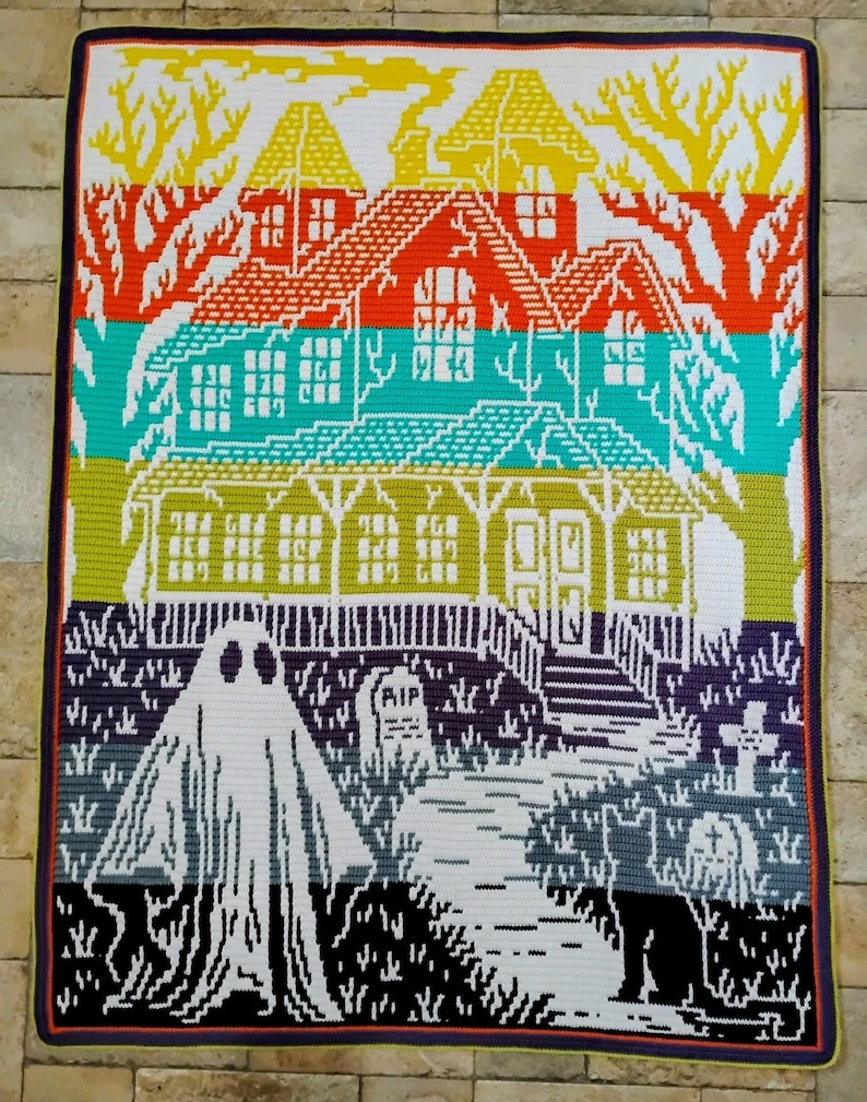 Op de afbeelding: Een kleurrijke gehaakte deken met een spookhuis-sc&egrave;ne. Het ontwerp omvat een geest, grafstenen en een pad dat naar een huis leidt. De deken gebruikt een verscheidenheid aan kleuren, waaronder oranje, groen en paars, met een zwarte rand.
