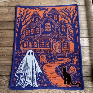 Op de afbeelding: Een Halloween-gehaakte deken met een spookhuis, een geest en een zwart katontwerp. De deken is voornamelijk blauw en oranje, met een gedetailleerde weergave van een spookachtige sc&egrave;ne. De geest is wit en de kat is zwart.