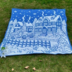 May include: A blue and white crocheted throw blanket with a winter scene of a house, trees, and a car. The house has a white roof and windows, and the trees are covered in snow. The car is driving down a snowy road.