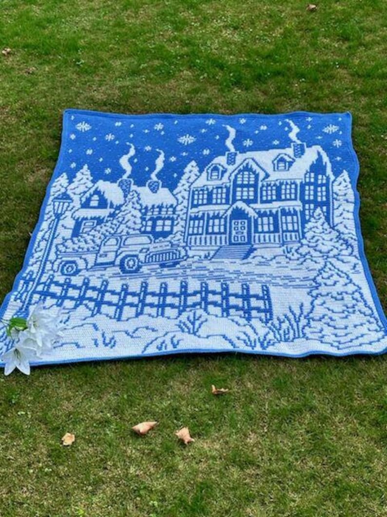 May include: A blue and white crocheted throw blanket with a winter scene of a house, trees, and a car. The house has a white roof and windows, and the trees are covered in snow. The car is driving down a snowy road.