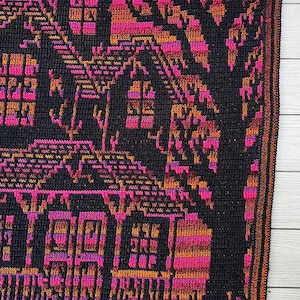 Op de afbeelding: Een gehaakte deken met een gedetailleerd ontwerp van huizen en een boom in zwart, roze en oranje. De deken heeft een donkere rand en wordt weergegeven op een licht houten oppervlak.