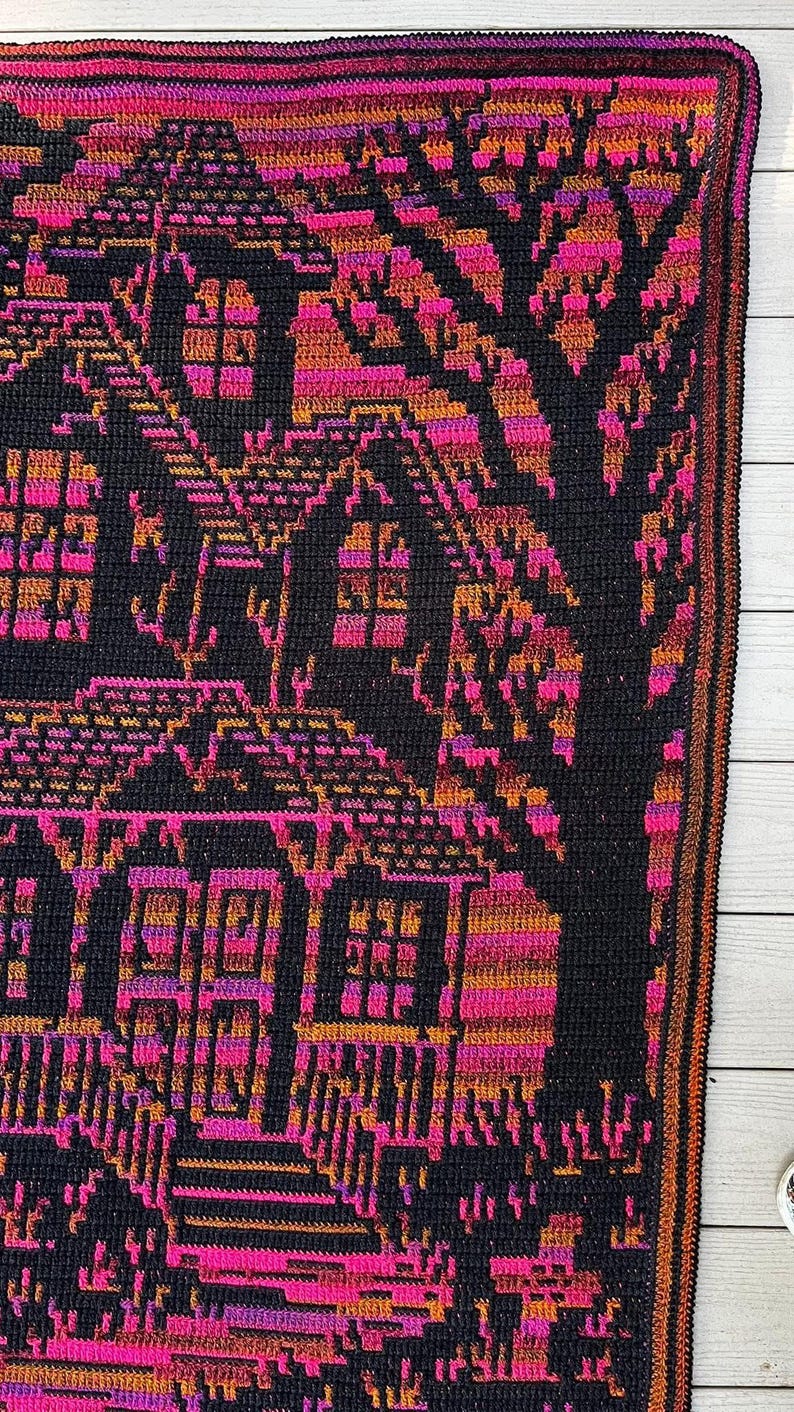 Op de afbeelding: Een gehaakte deken met een gedetailleerd ontwerp van huizen en een boom in zwart, roze en oranje. De deken heeft een donkere rand en wordt weergegeven op een licht houten oppervlak.