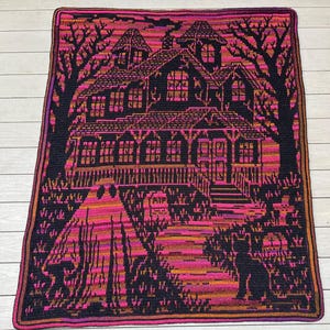 Op de afbeelding: Een gehaakte deken met een spookachtig spookhuis. Het ontwerp omvat een huis, bomen, een geest en een zwarte kat, allemaal in tinten zwart, roze, oranje en paars. De deken heeft een donkere rand.